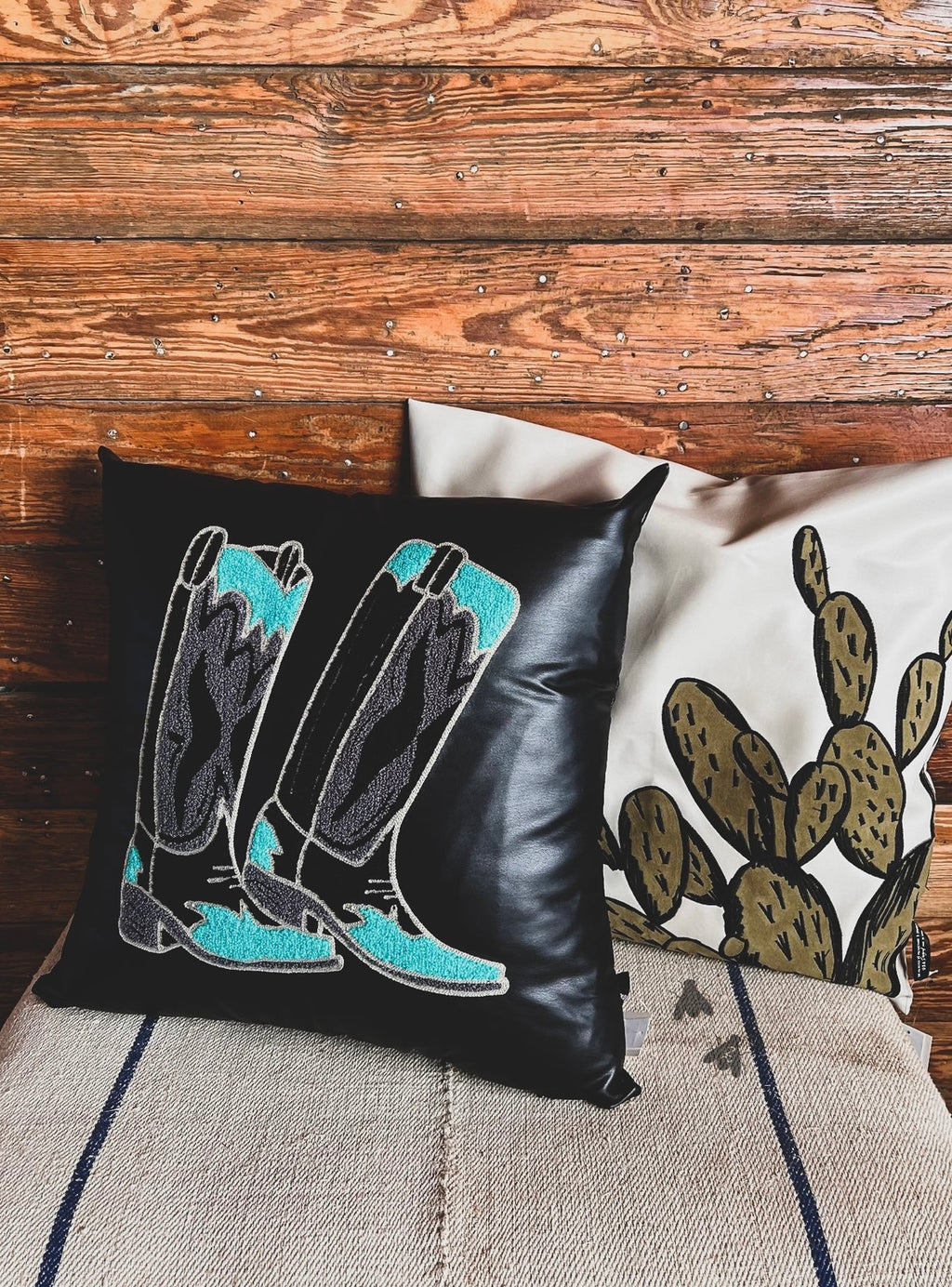 A decorative pillow case with a black background featuring an embroidered design of boots in teal and grey colors. 