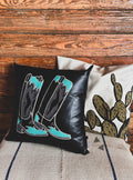 A decorative pillow case with a black background featuring an embroidered design of boots in teal and grey colors. 