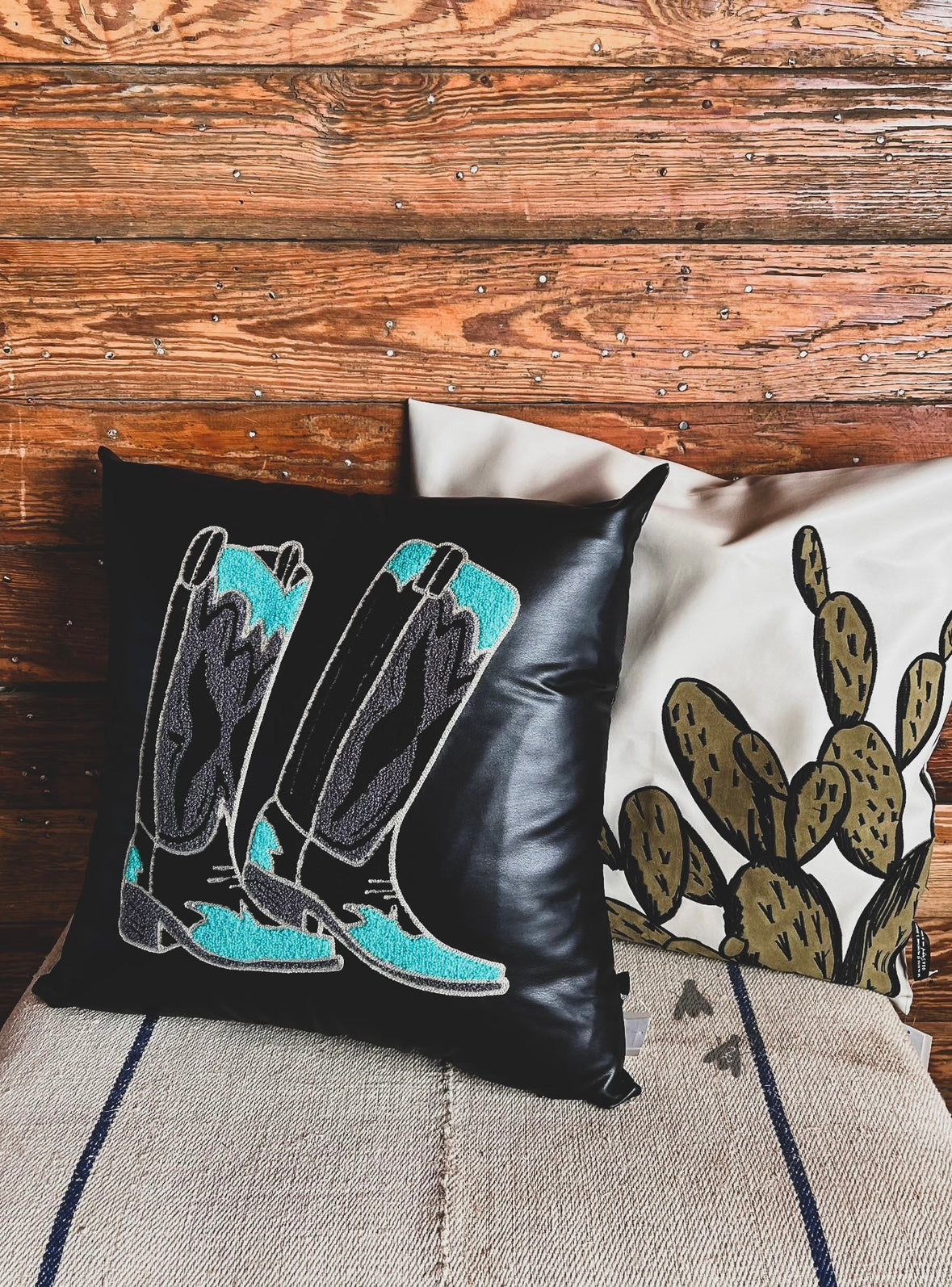 A decorative pillow case with a black background featuring an embroidered design of boots in teal and grey colors. 